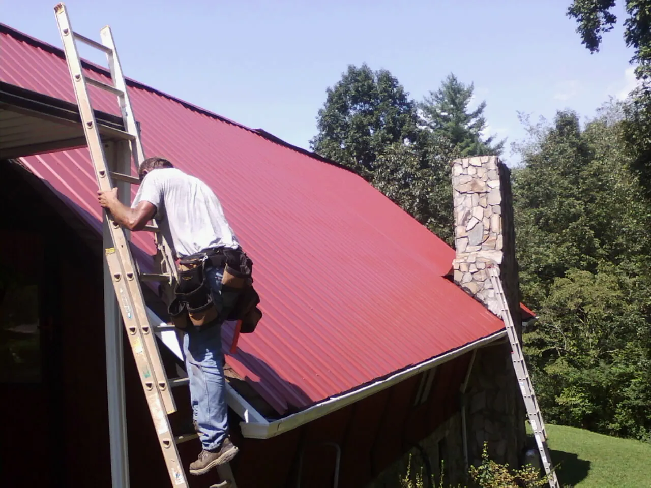 Roofer on a ladder beside a red metal roof and stone chimney — Soffit & Fascia Repair by Roofing Brothers in Arbovale